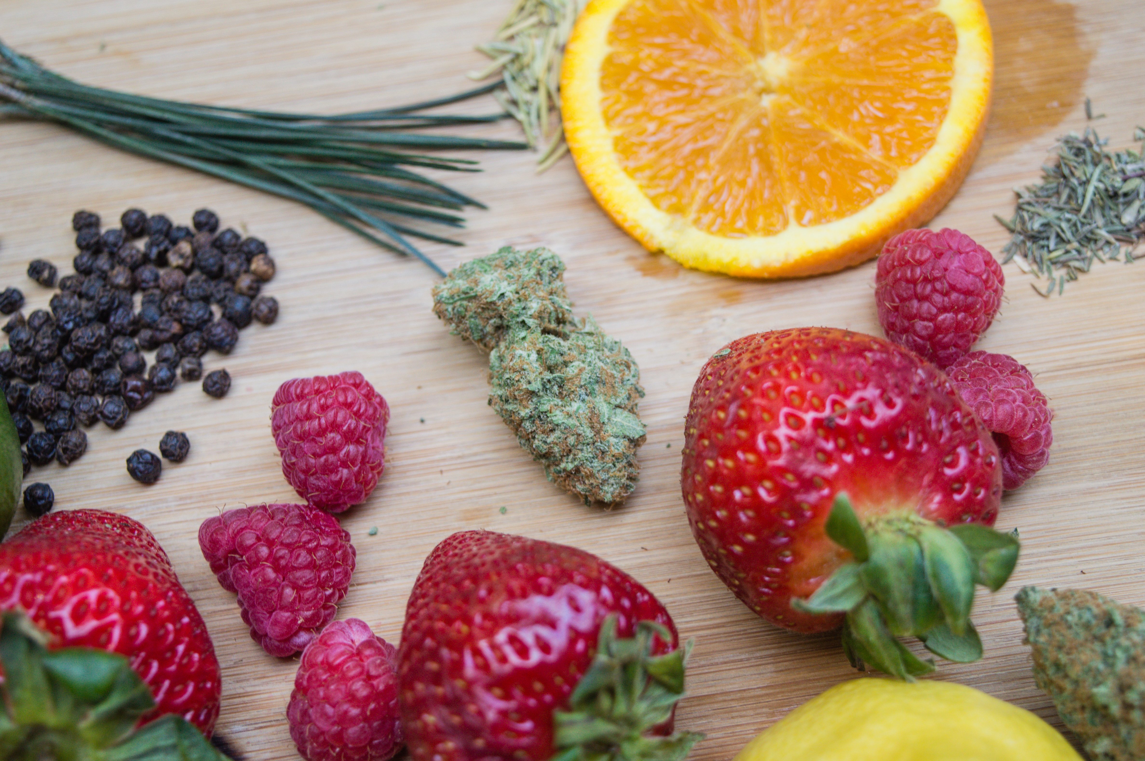 A Cannabis bud sits on a cutting board surrounded by strawberries, diced orange, raspberries, peppercorn and pine.